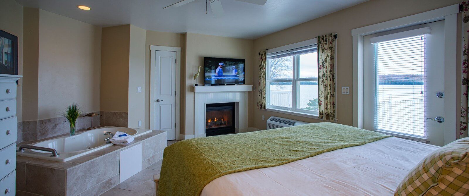 Bedroom with bed, jacuzzi tub, gas fireplace with TV and large windows overlooking water