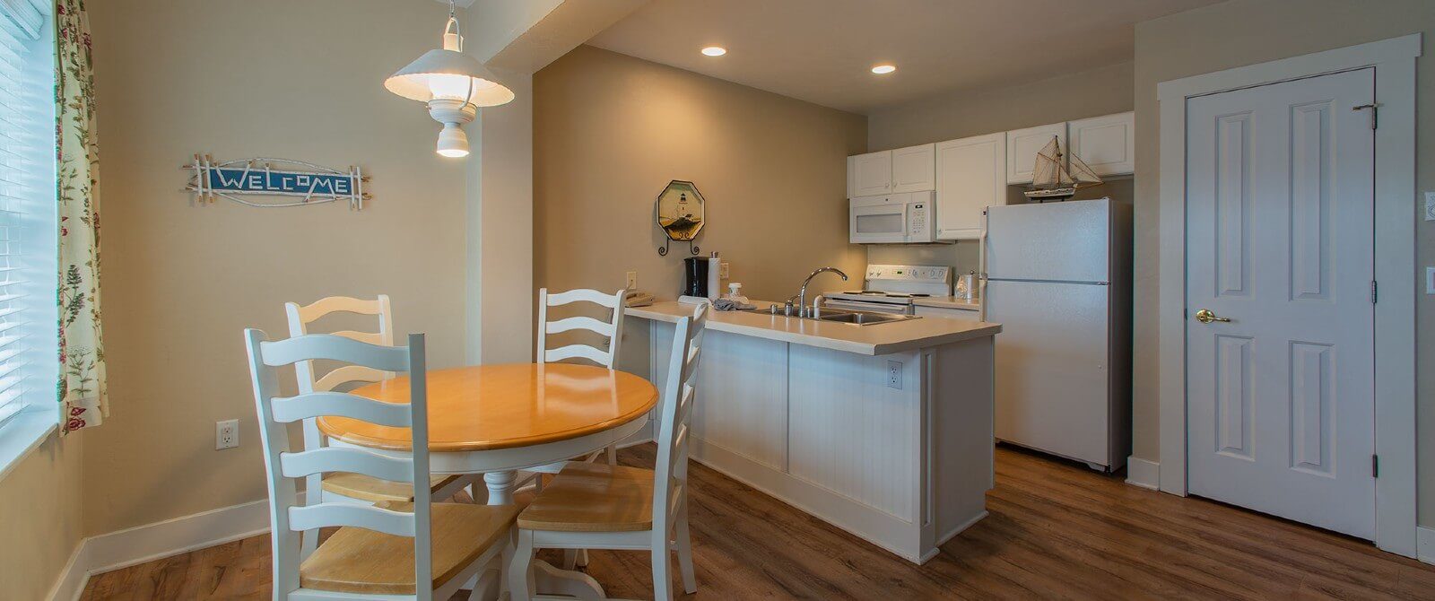 Kitchenette area with round wood table and four white chairs by a window with floral curtains