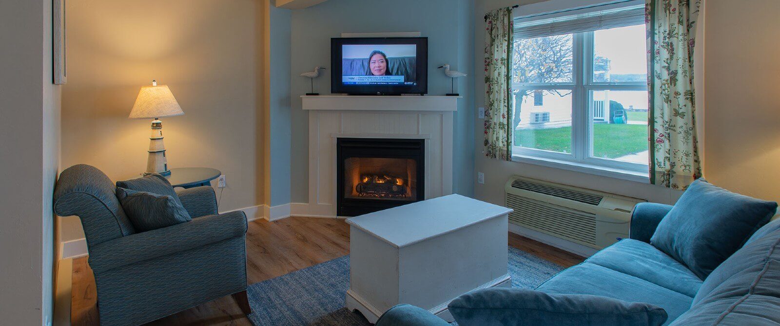 Living room with blue couch and chair, white table, TV over a gas fireplace and large window with curtains