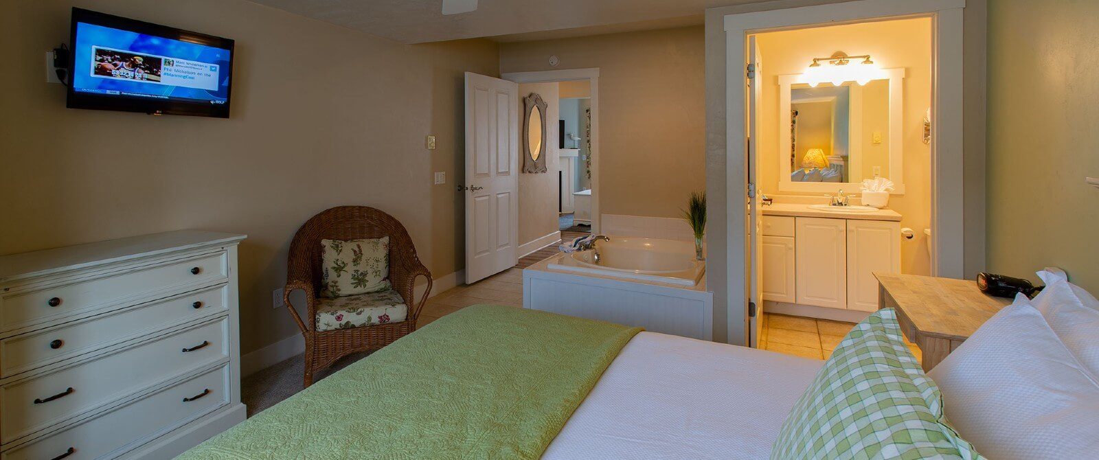 Bedroom with white and green bed, dresser, TV, jacuzzi tub and doorway open to a bathroom