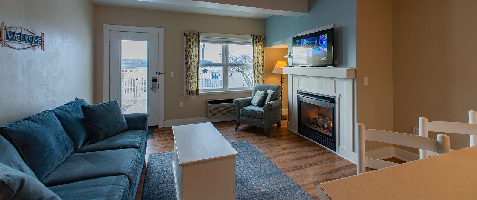 Living room with blue couch and chair, white table, TV over a gas fireplace and doorway to a patio