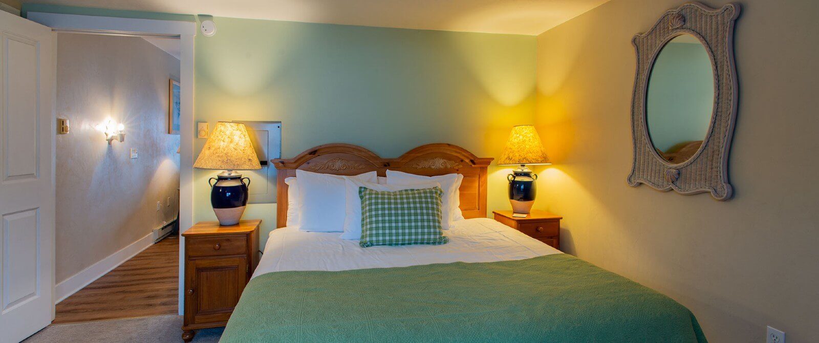 Small bedroom with queen bed in green and white, framed mirror and open door to a hallway