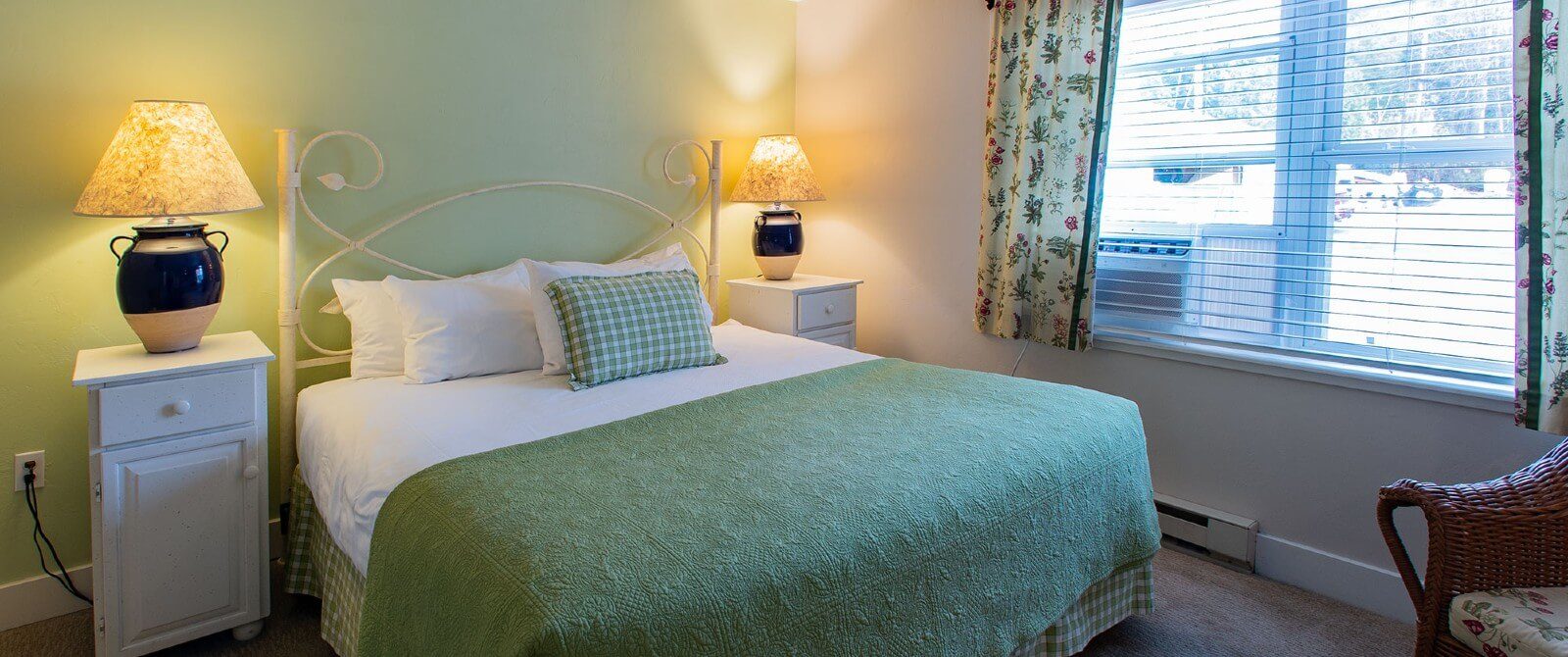 Small bedroom with bed in green and white, two side tables with lamps and large window with blinds