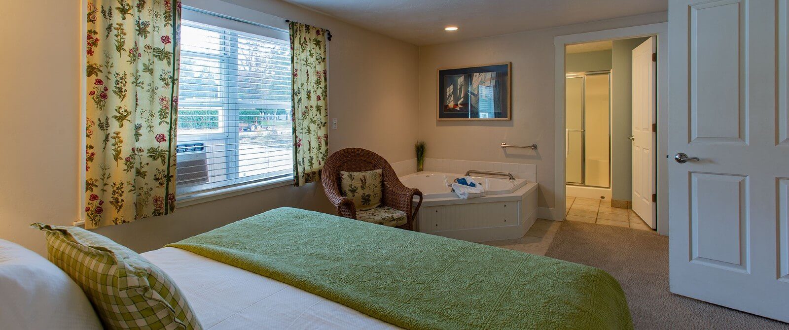 Small bedroom with bed in white and green linens, corner jacuzzi tub, window with blinds and doorway open to a bathroom