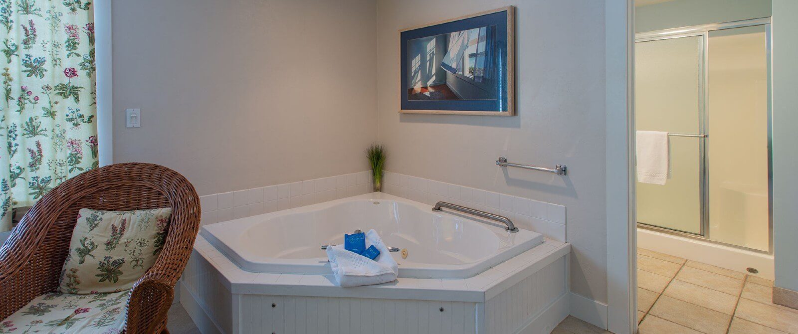 Corner jacuzzi tub, wicker sitting chair and doorway into a bathroom with a stand up shower with glass doors