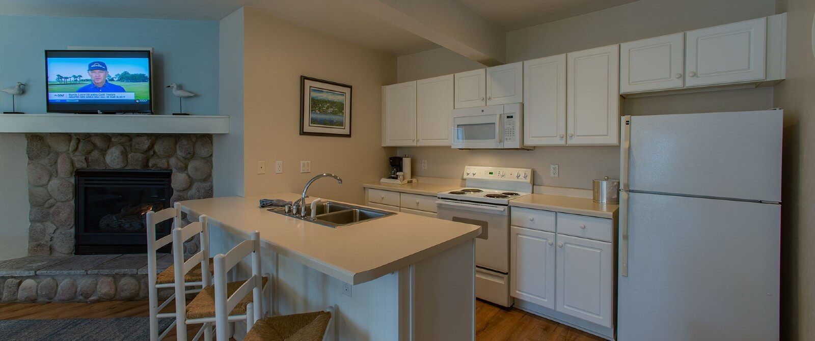 Kitchen with white cabinets, island countertop with sink and three stools, next to a stone fireplace