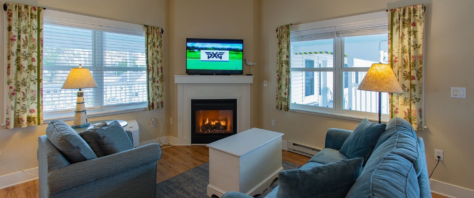 Living room with plush blue couch and chair, coffee table, gas fireplace and large windows with blinds and floral curtains