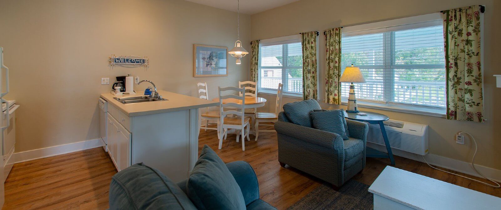 Large room with kitchenette, table with four chairs, blue couch and chair by two large windows with blinds and floral curtains