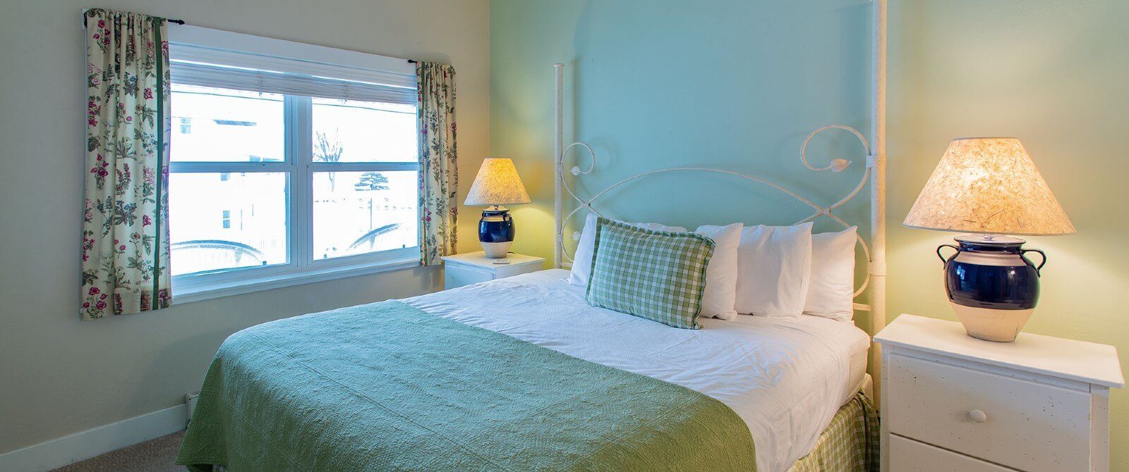 Small bedroom with bed in green and white linens, two side tables with lamps and large window with floral curtains