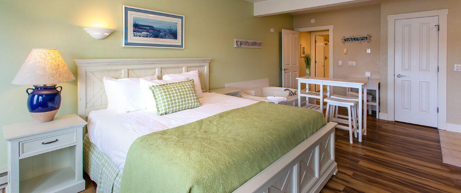 Bright and spacious bedroom with king bed in green and white linens, soaker tub, bistro table with stools and hardwood floors