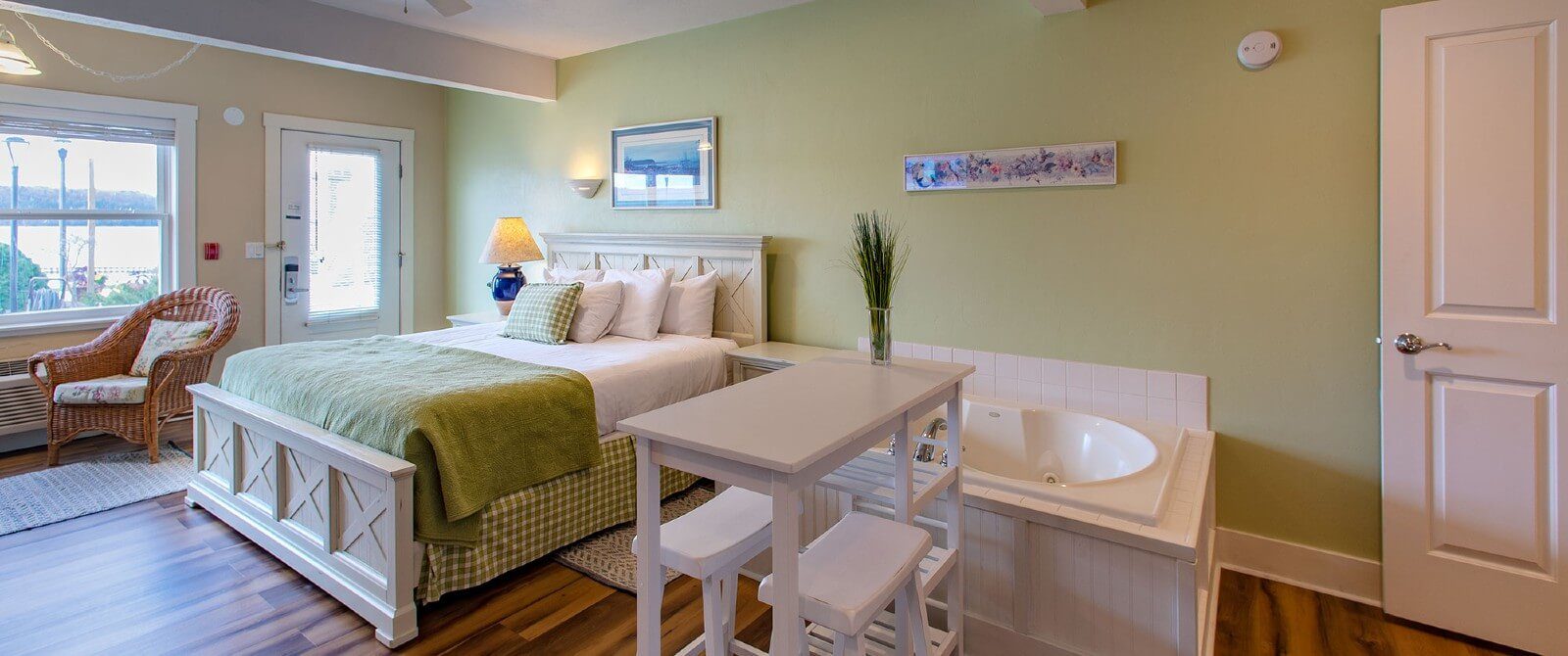 Bright and spacious bedroom with king bed in green and white linens, soaker tub, bistro table with stools and hardwood floors