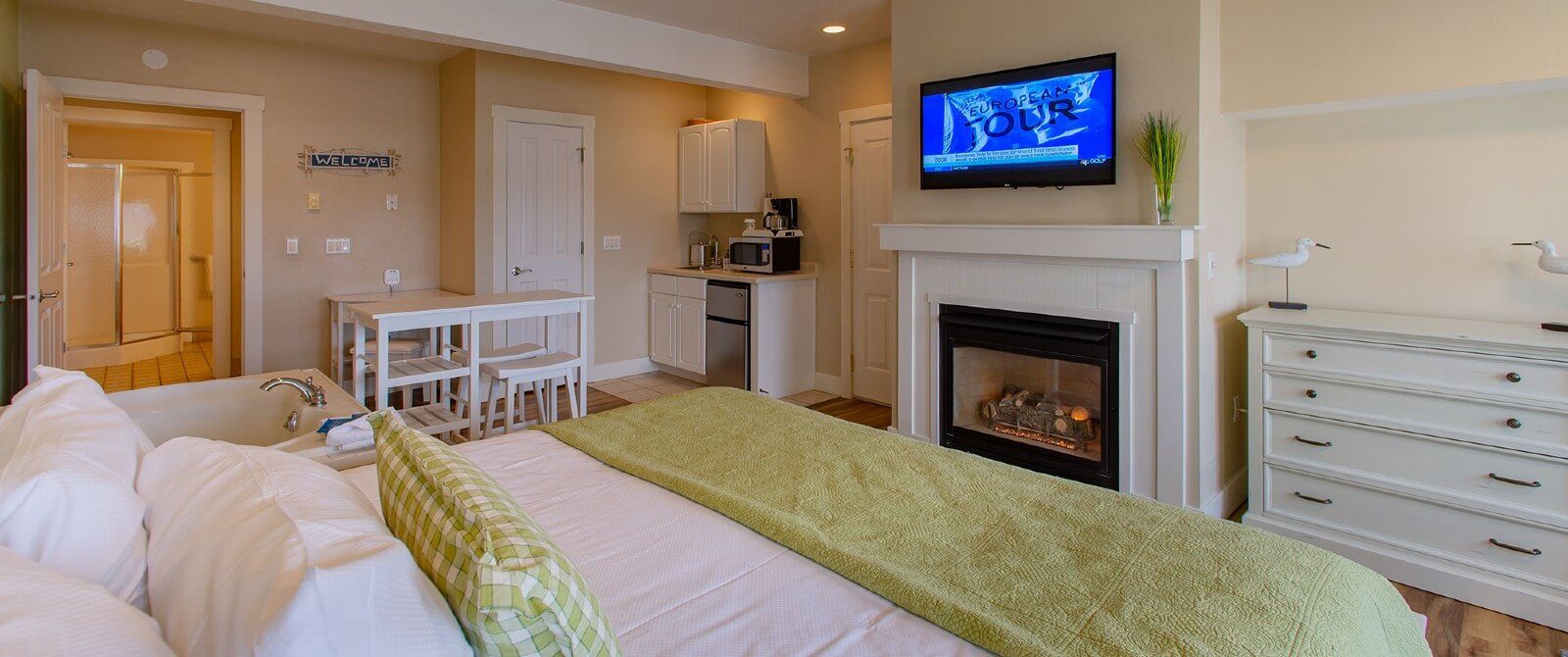 Bright and spacious bedroom with king bed in green and white linens, soaker tub, bistro table with stools, gas fireplace and kitchenette