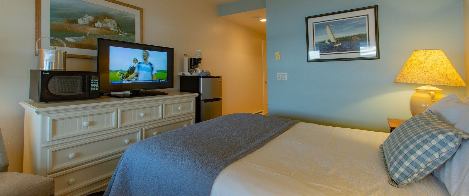 Bedroom with queen bed, blue wall with framed artwork, dresser with TV and microwave