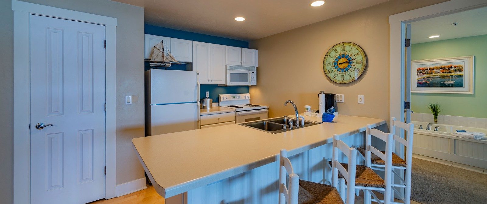 Full kitchen with white cabinets, island sink, three barstools and doorway into a room with a jacuzzi