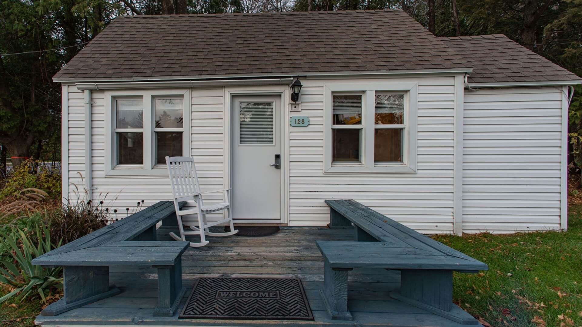 Small white cottage with four windows, front deck with bench seats and one white rocking chair