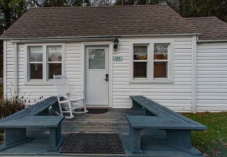 Small white cottage with four windows, front deck with bench seats and one white rocking chair