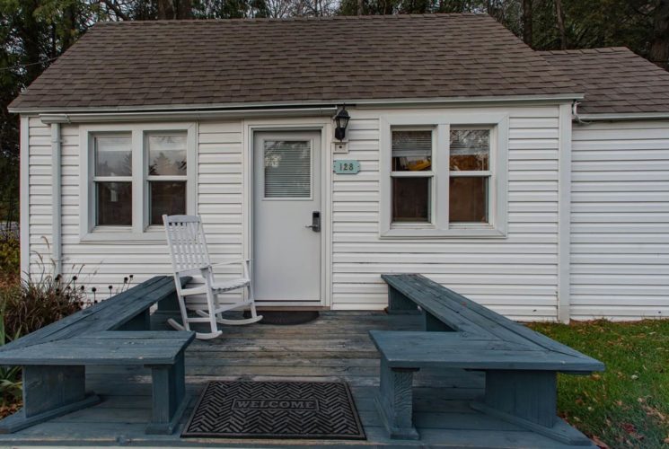 Small white cottage with four windows, front deck with bench seats and one white rocking chair