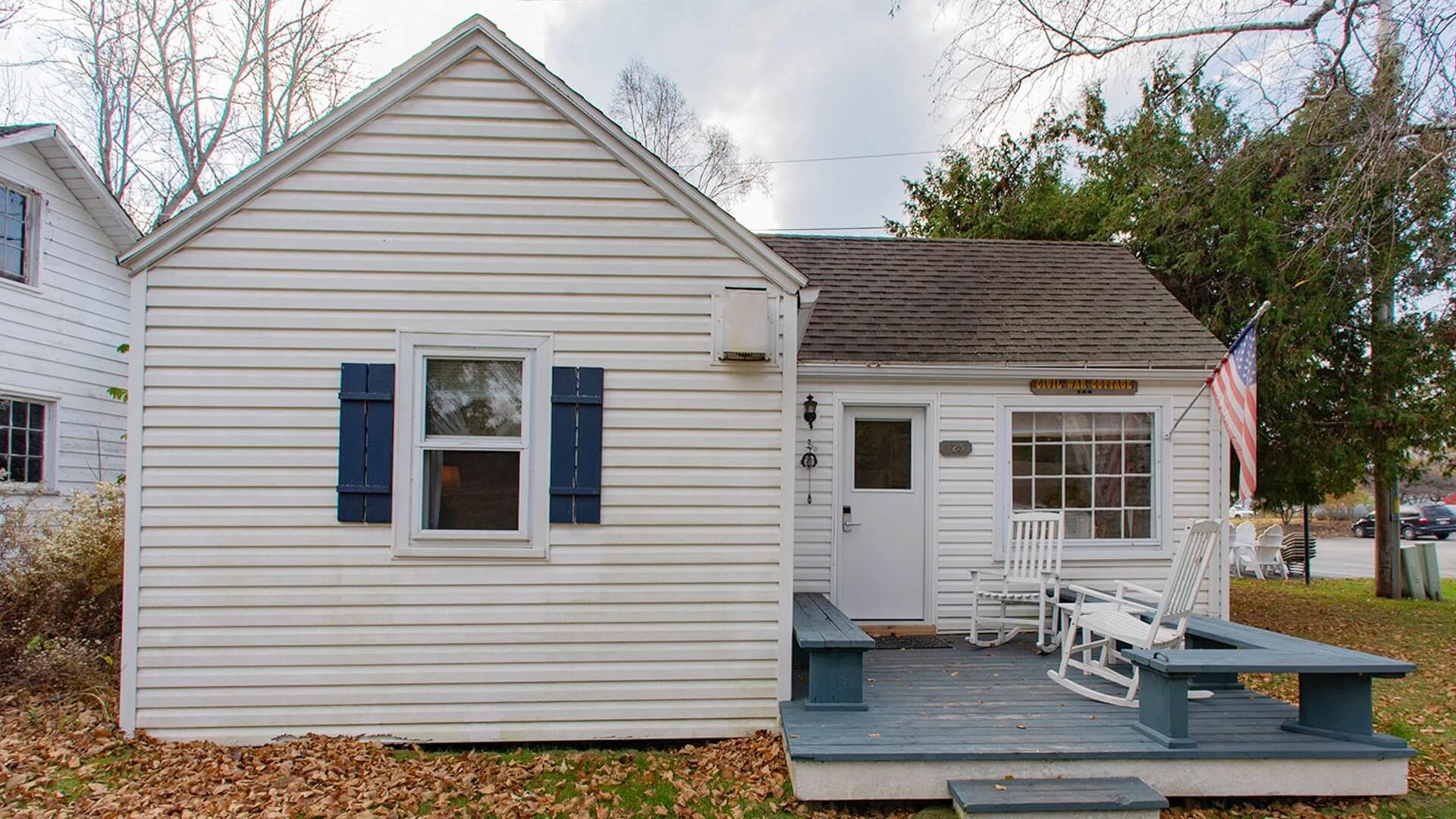 Exterior of a small white cottage with front deck with bench seats, two white rocking chairs and an American flag on a pole by front window