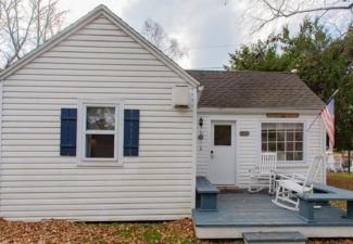 Exterior of a small white cottage with front deck with bench seats, two white rocking chairs and an American flag on a pole by front window