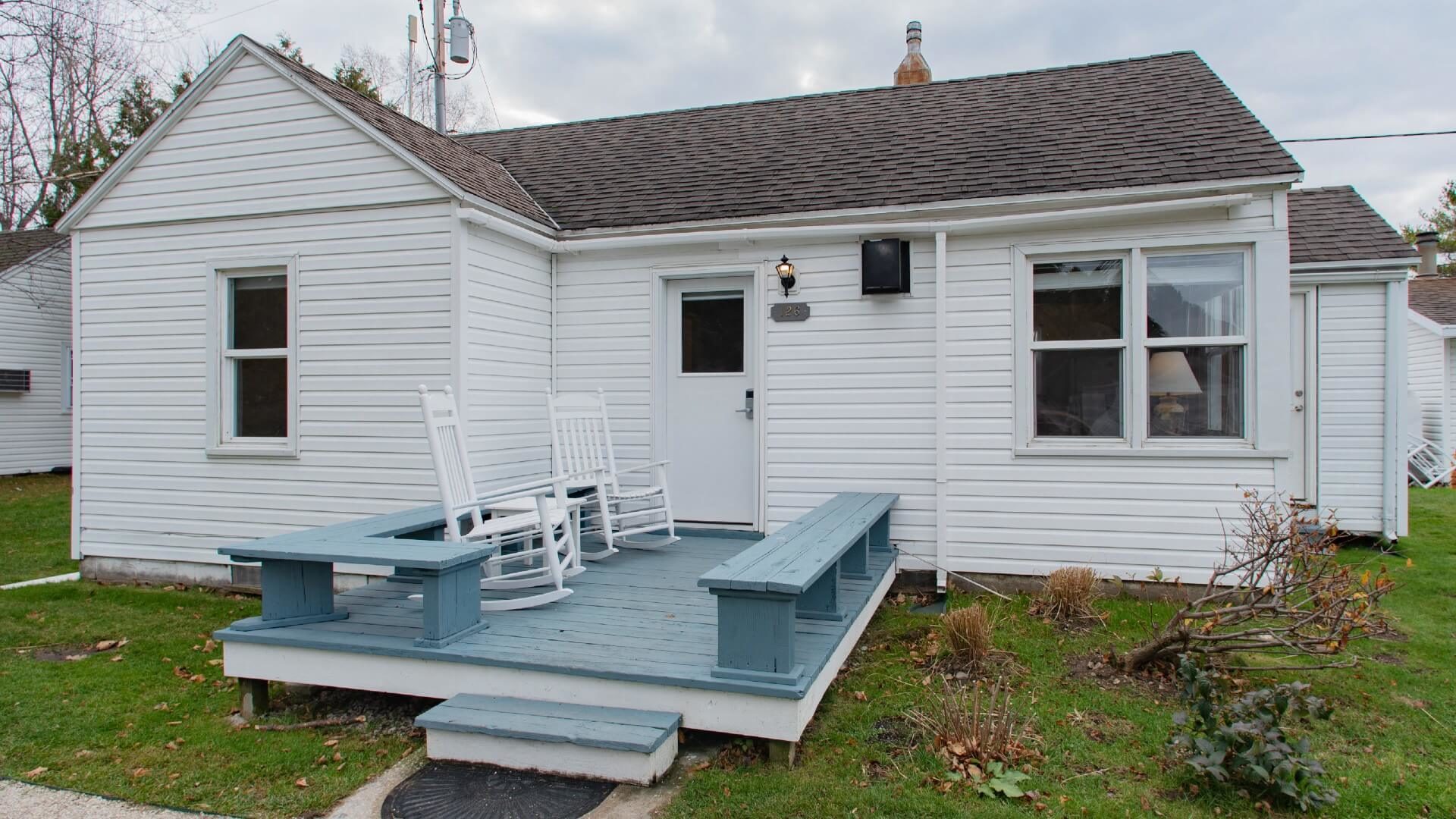 Small white cottage with front deck with bench seats, two white rocking chairs and small plot of grass