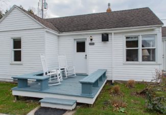 Small white cottage with front deck with bench seats, two white rocking chairs and small plot of grass
