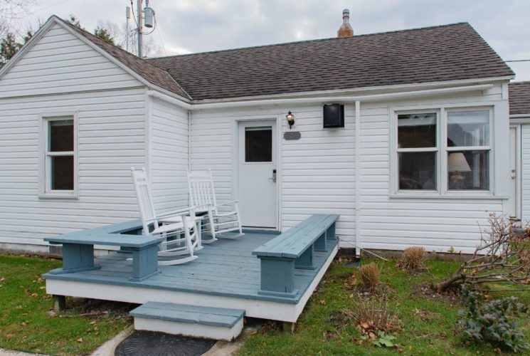 Small white cottage with front deck with bench seats, two white rocking chairs and small plot of grass