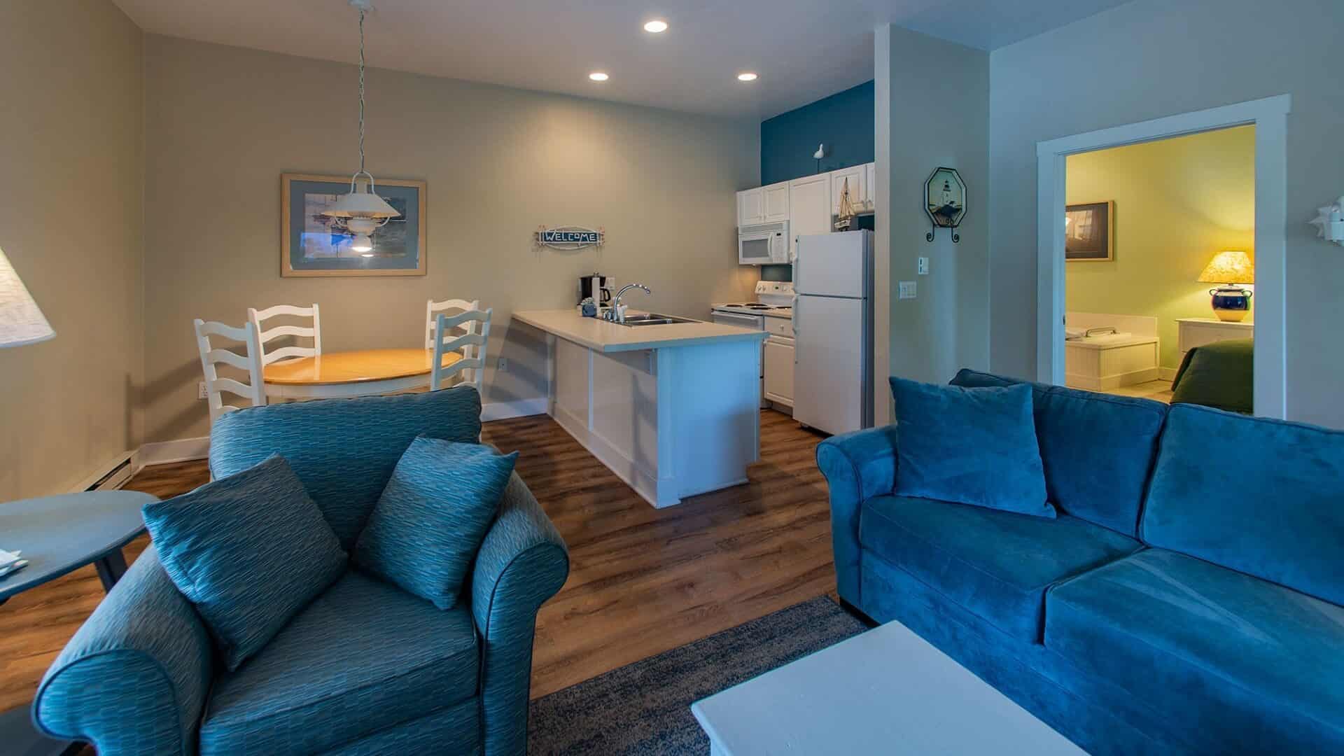 Hotel suite with kitchenette, hardwood floors, blue couch and chair and doorway into a bedroom with a jacuzzi tub