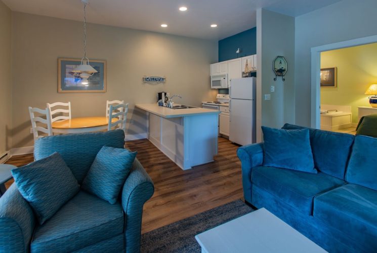 Hotel suite with kitchenette, hardwood floors, blue couch and chair and doorway into a bedroom with a jacuzzi tub