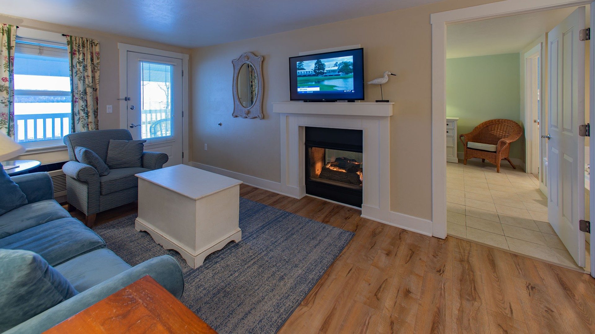 Living room with couch, chair and white coffee table in front of a gas fireplace with doorway into a bedroom