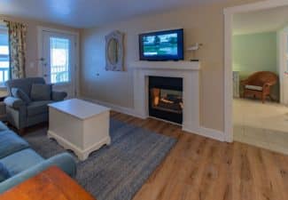 Living room with couch, chair and white coffee table in front of a gas fireplace with doorway into a bedroom