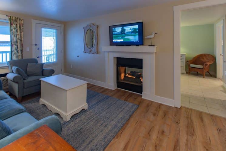 Living room with couch, chair and white coffee table in front of a gas fireplace with doorway into a bedroom