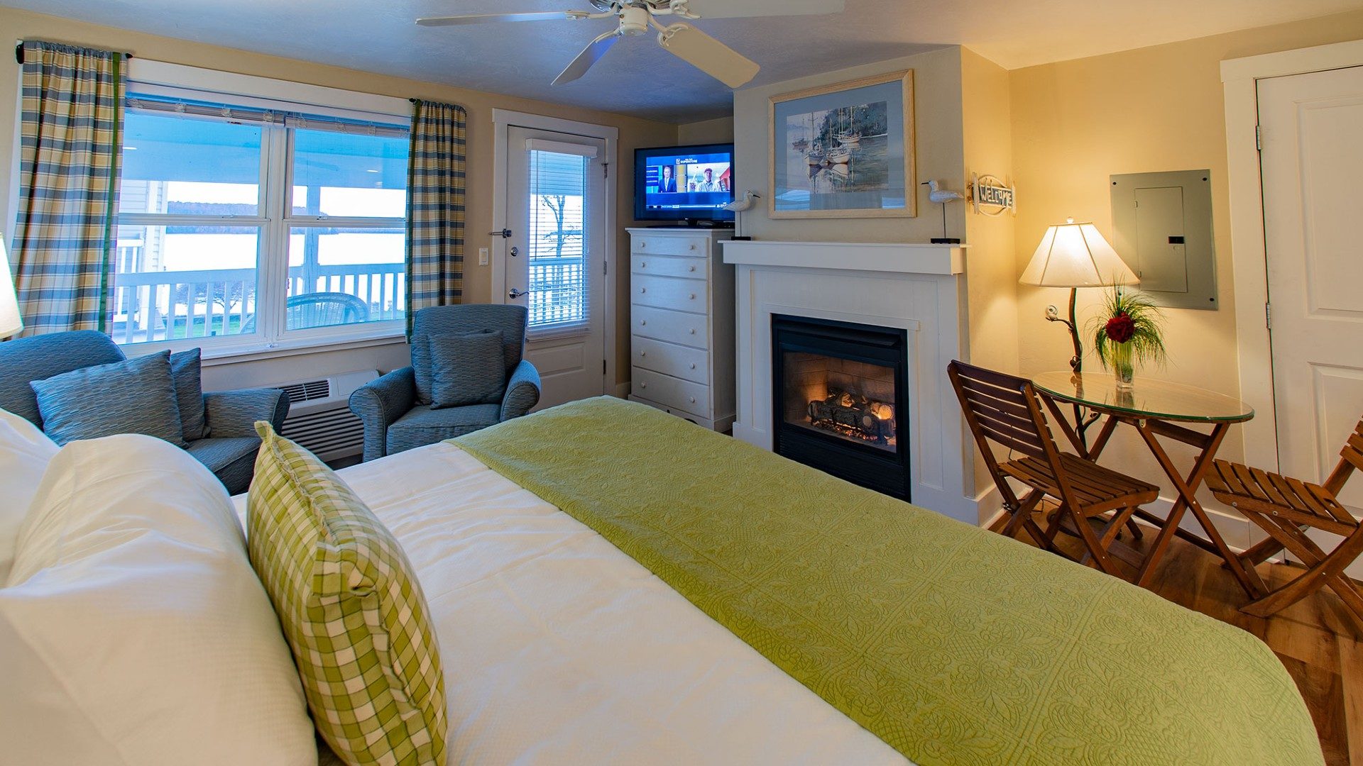 Elegant bedroom with king bed, sitting chairs, gas fireplace, dresser with TV and large window overlooking the water