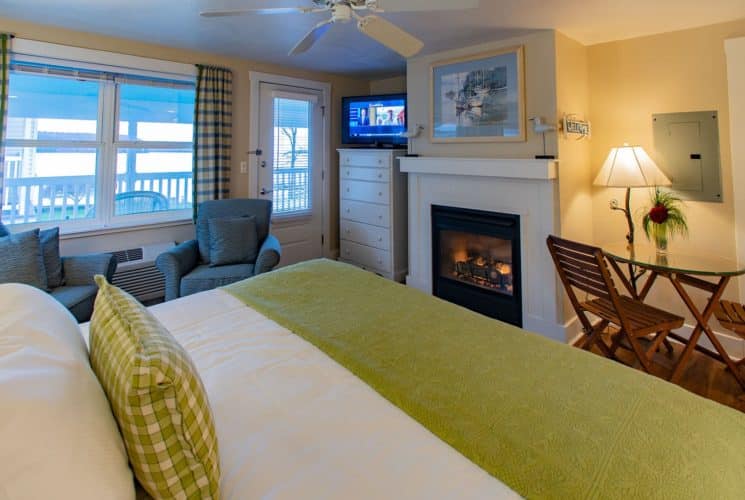 Elegant bedroom with king bed, sitting chairs, gas fireplace, dresser with TV and large window overlooking the water