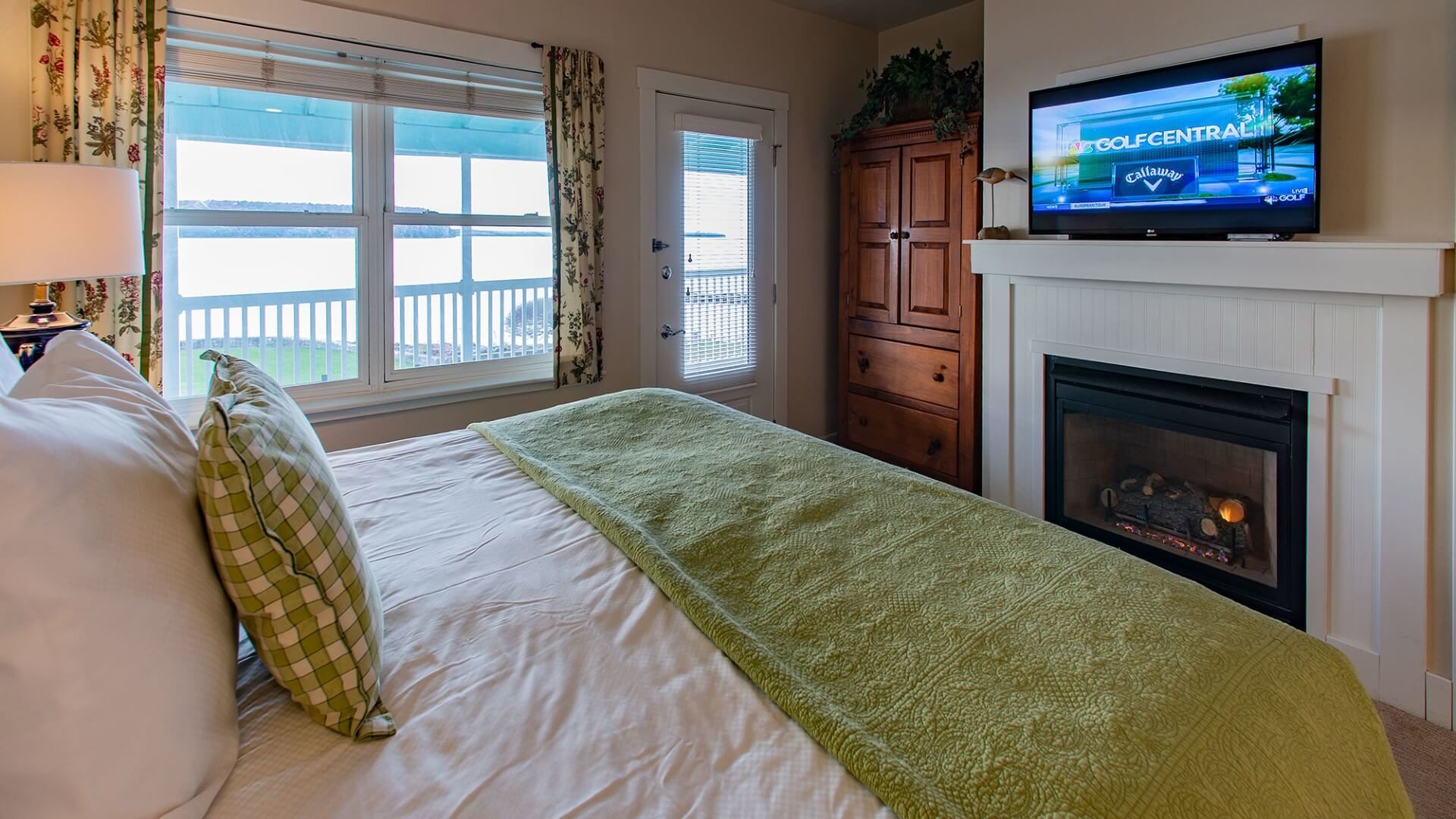 Cozy bedroom with bed in white and green linens, TV above gas fireplace, large armoire and window with floral curtains