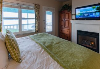 Cozy bedroom with bed in white and green linens, TV above gas fireplace, large armoire and window with floral curtains