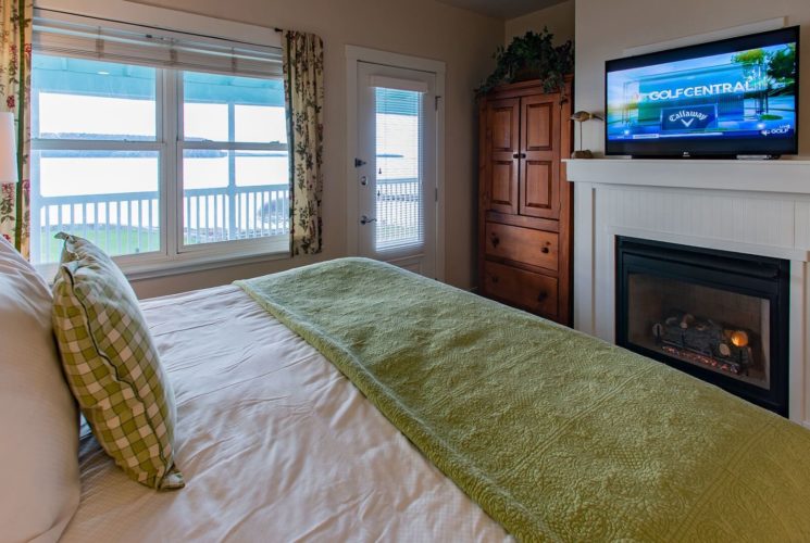 Cozy bedroom with bed in white and green linens, TV above gas fireplace, large armoire and window with floral curtains