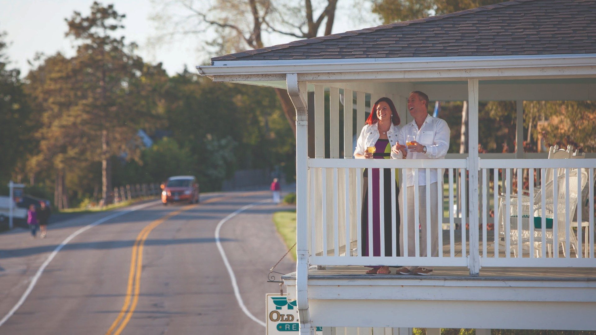 A man and woman standing on an upper deck holding wine glasses overlooking a two lane road on a sunny day