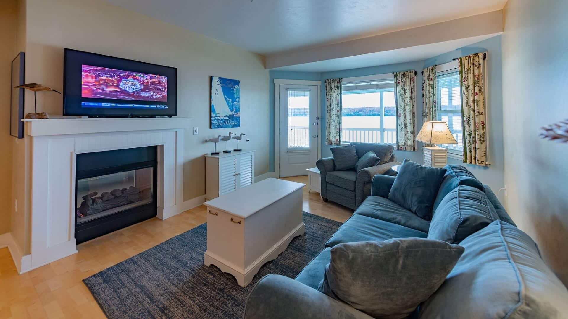 Living room with large couch, chair, coffee table, lamp, gas fireplace with TV above and bay window overlooking the water
