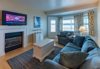 Living room with large couch, chair, coffee table, lamp, gas fireplace with TV above and bay window overlooking the water