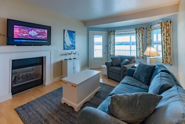 Living room with large couch, chair, coffee table, lamp, gas fireplace with TV above and bay window overlooking the water