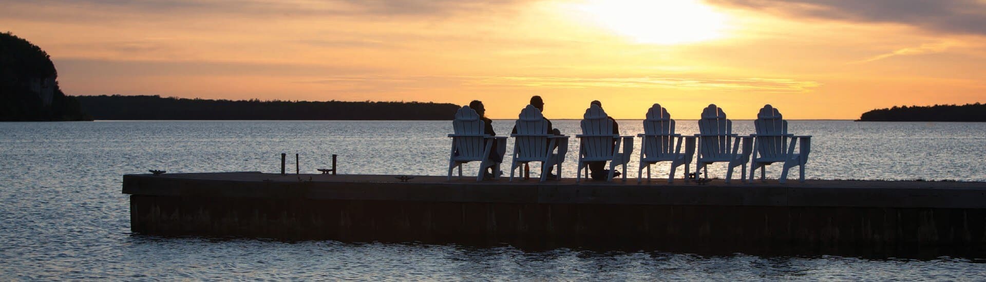 Six Adirondack chairs at the edge of a dock overlooking a body of water at sunset