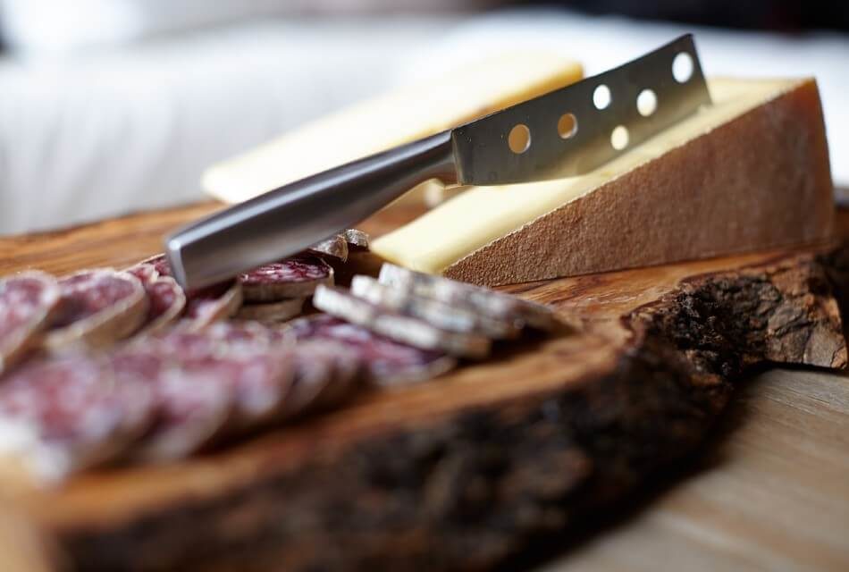 A thick wood platter with two blocks of cheese, a knife and slices of summer sausage