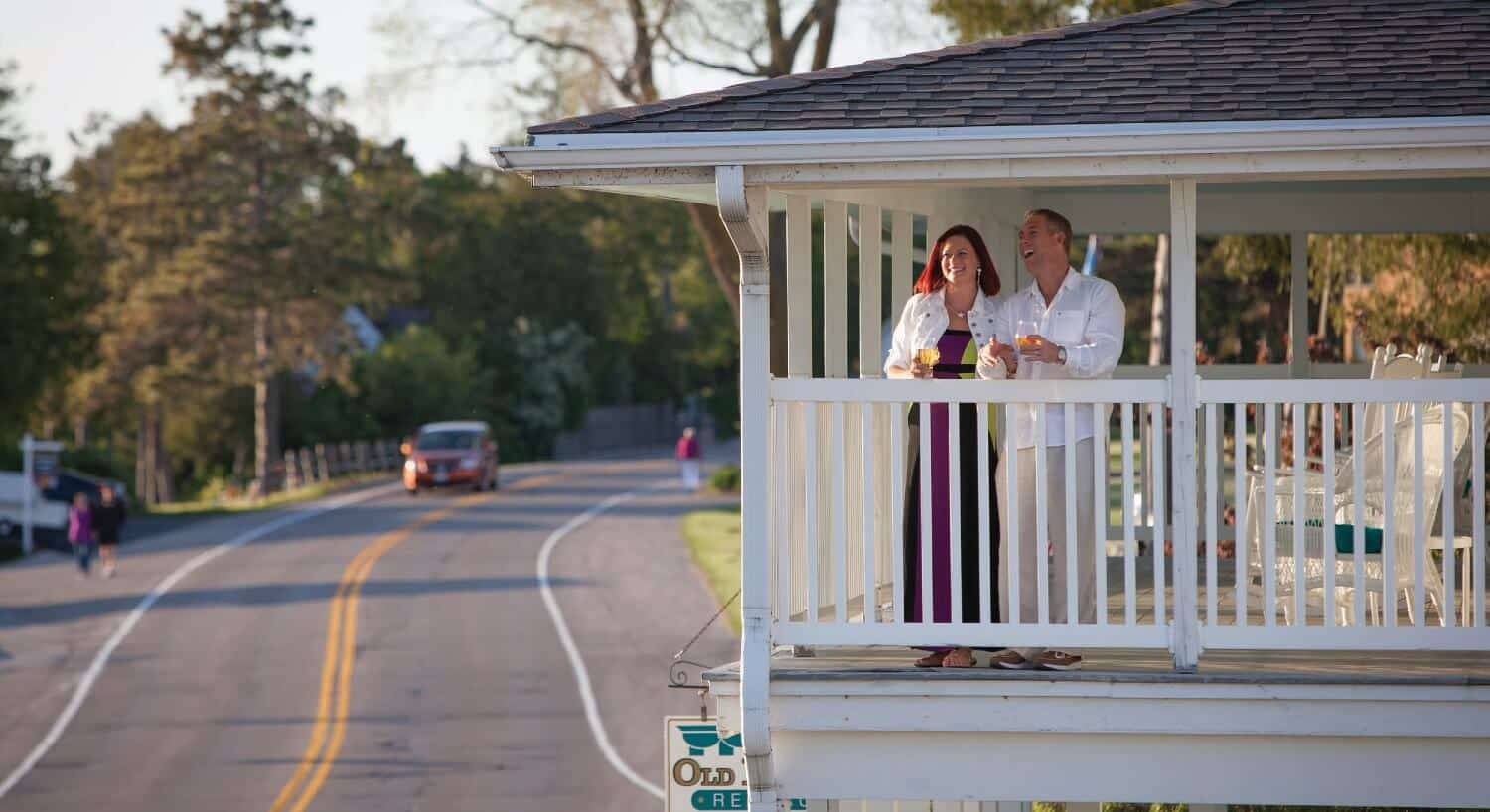 A man and woman standing on an upper level deck with white railings overlooking a two lane road with trees