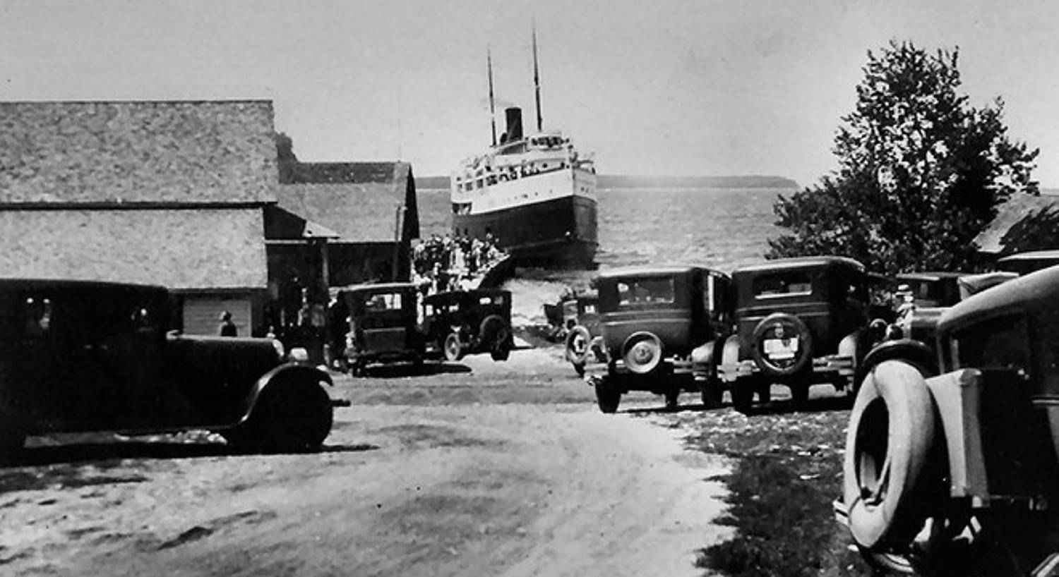 A black and white historic photograph of a large ship coming in to dock at a small town with antique cars