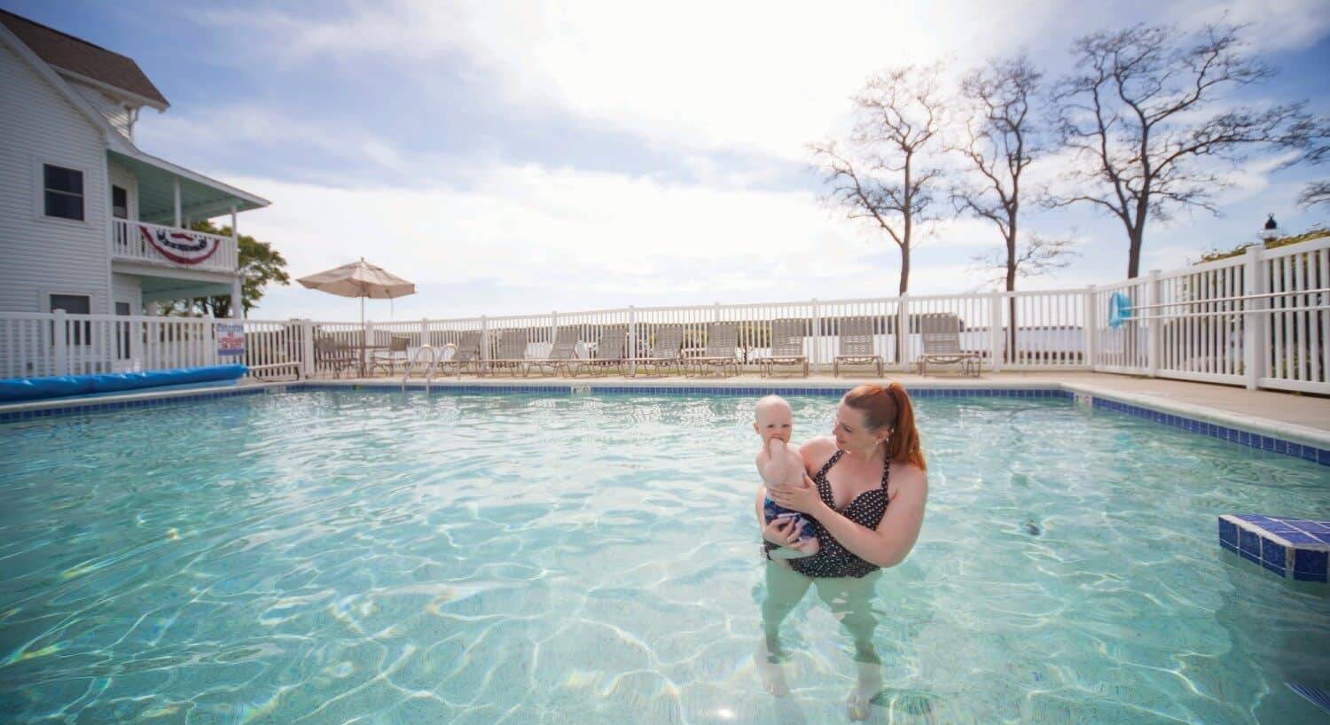 A woman bolding a baby in a large outdoor pool next to a hotel building and overlooking the water