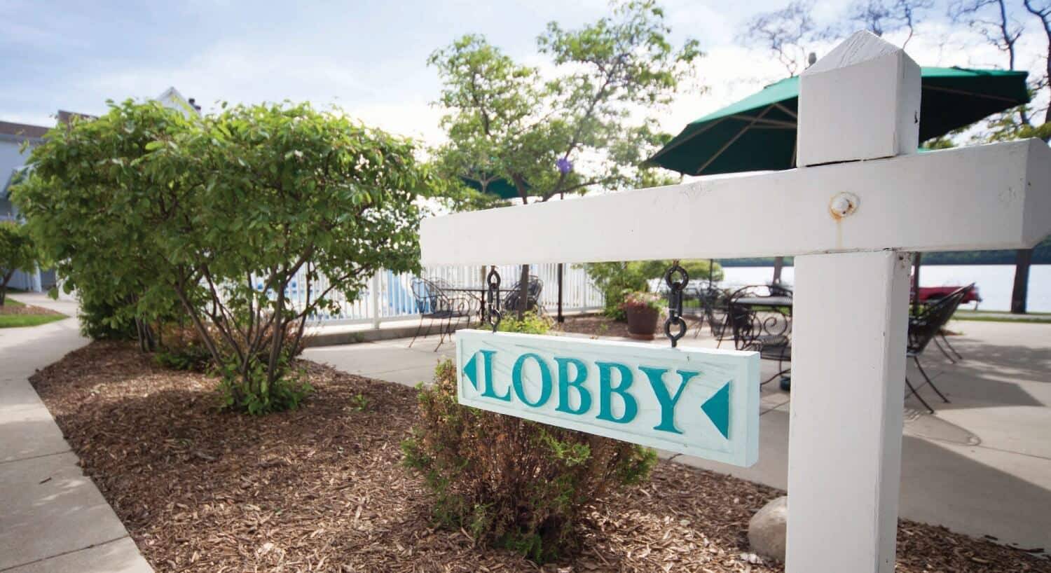 Sign pointing towards a lobby on a white post near trees and an outdoor patio table