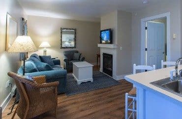 Living room with couch, chairs, coffee table, gas fireplace with flat screen TV, next to a small kitchen