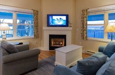 Living room with plush couch, chair, table on a blue rug, fireplace with TV and two large windows with floral curtains