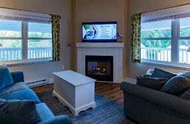 Living room with plush couch and chair, white table, gas fireplace with flat screen TV, two large picture windows with curtains