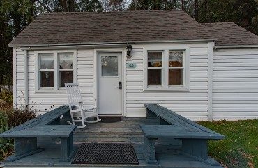 Small white cottage with front deck, bench seats and one white rocking chair